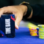 Mão de jogador segurando cartões de time bank ao lado das fichas em uma mesa de torneio de poker