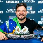 Fabiano Kovalski sorri com a bandeira do Brasil no ombro, cartas QQ, fichas e troféu Mystery Bounty do PokerStars à frente