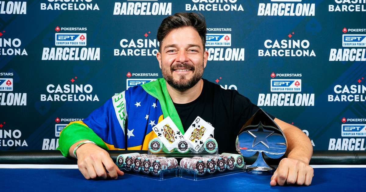 Fabiano Kovalski sorri com a bandeira do Brasil no ombro, cartas QQ, fichas e troféu Mystery Bounty do PokerStars à frente