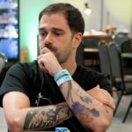 Felipe Boianovsky, de camiseta preta, apoia o queixo na mão e cruza os braços tatuados sobre a mesa de um torneio do BSOP