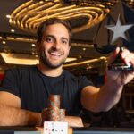 João Simão sorri e mostra o troféu de campeão do NAPT Las Vegas, em formato de naipe de espadas com a estrela do PokerStars, sentado à mesa com fichas