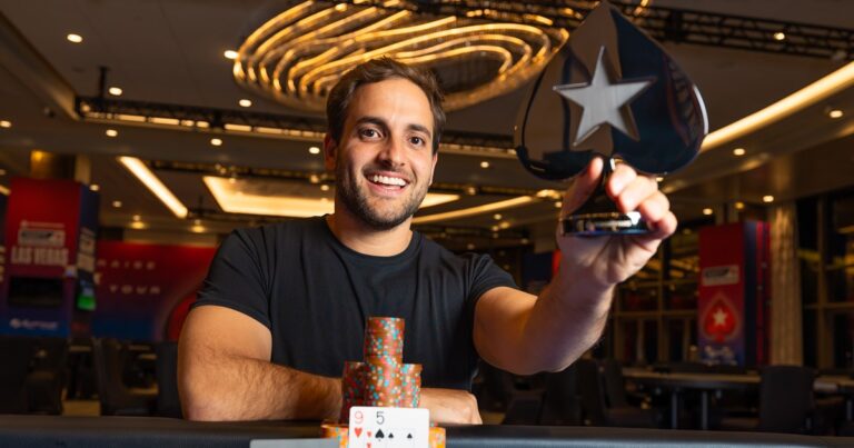 João Simão sorri e mostra o troféu de campeão do NAPT Las Vegas, em formato de naipe de espadas com a estrela do PokerStars, sentado à mesa com fichas