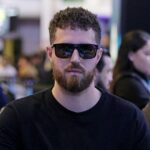 Luan Barbosa, de óculos escuros e camiseta preta, sentado a uma mesa de torneio ao vivo do BSOP, com outros jogadores desfocados ao fundo