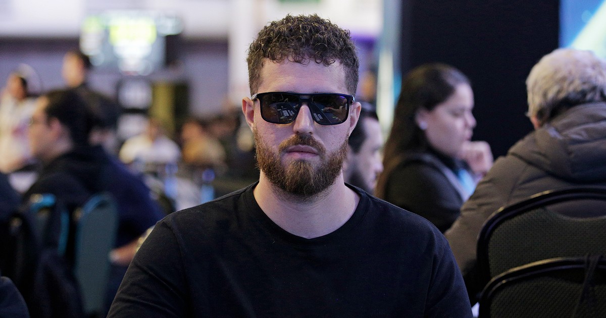 Luan Barbosa, de óculos escuros e camiseta preta, sentado a uma mesa de torneio ao vivo do BSOP, com outros jogadores desfocados ao fundo