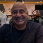 Wendel Cader, careca e de camiseta azul-escura, sorri sentado a uma mesa de torneio do JPT, com o salão e outros jogadores desfocados ao fundo