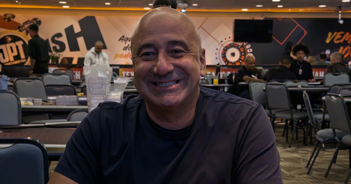 Wendel Cader, careca e de camiseta azul-escura, sorri sentado a uma mesa de torneio do JPT, com o salão e outros jogadores desfocados ao fundo